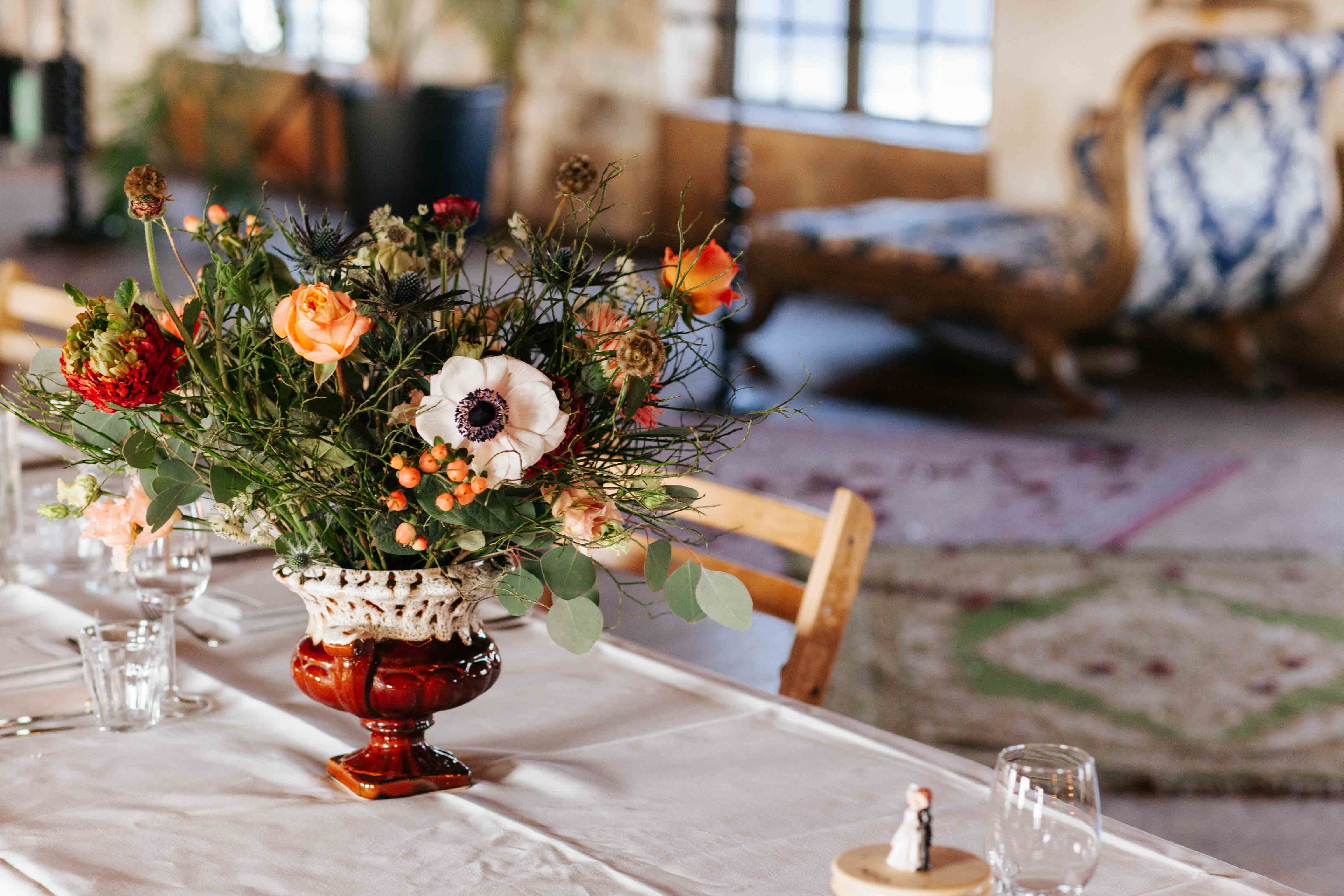 Een mooi gestileerde tafel, met daarop een oude vaas met een kleurrijk boeket bloemen. De bloemen zijn oranje, rood en wit, met veel groen tussendoor. Op de achtergrond zijn een lage bank en twee gekleurde vloerkleden zichtbaar.