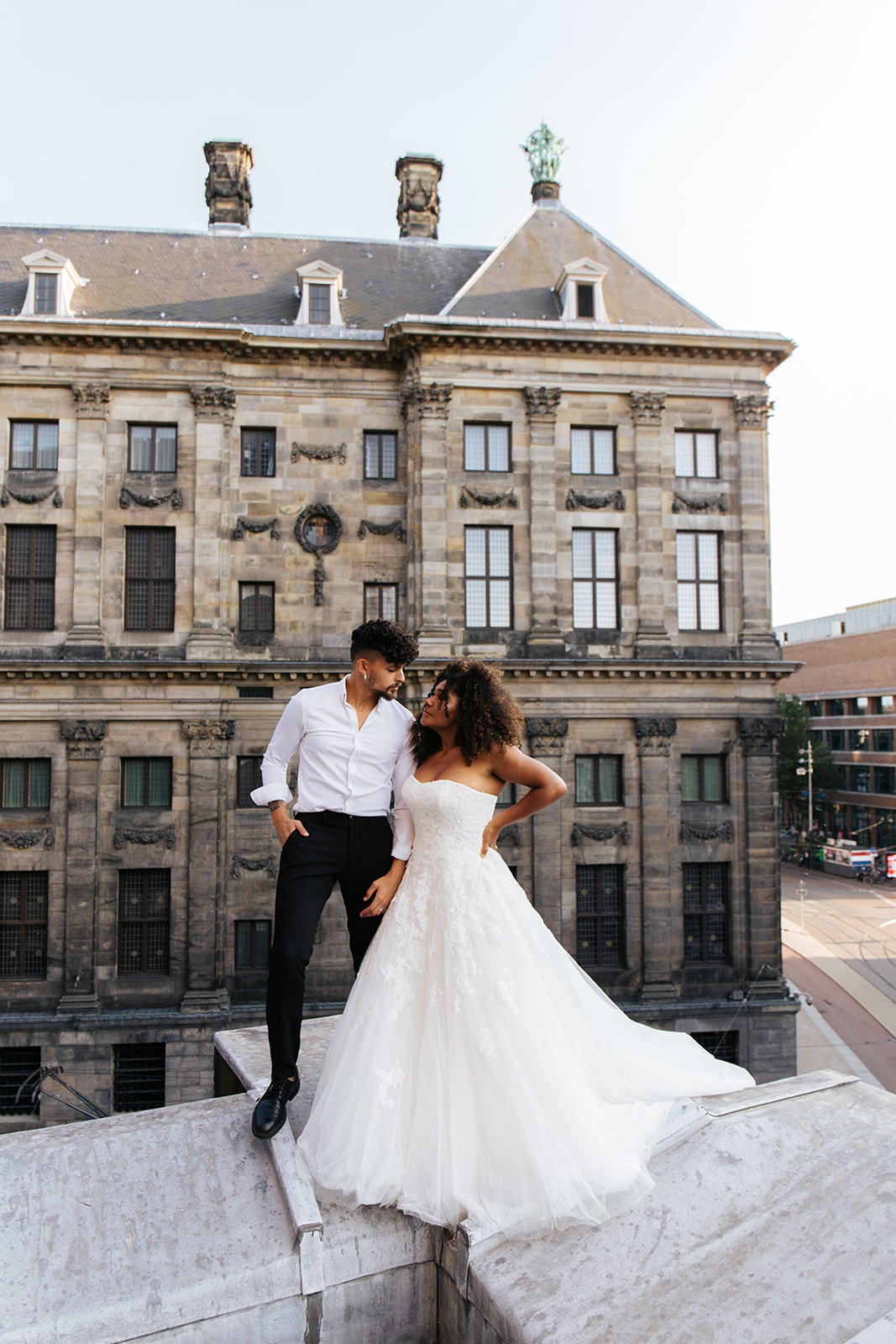 Een bruidspaar dat boven op het dak van de nieuwe kerk staat, in Amsterdam. Ze poseren op het randje van het dak, met uitzicht op de straten en de omliggende gebouwen van Centrum Amsterdam.