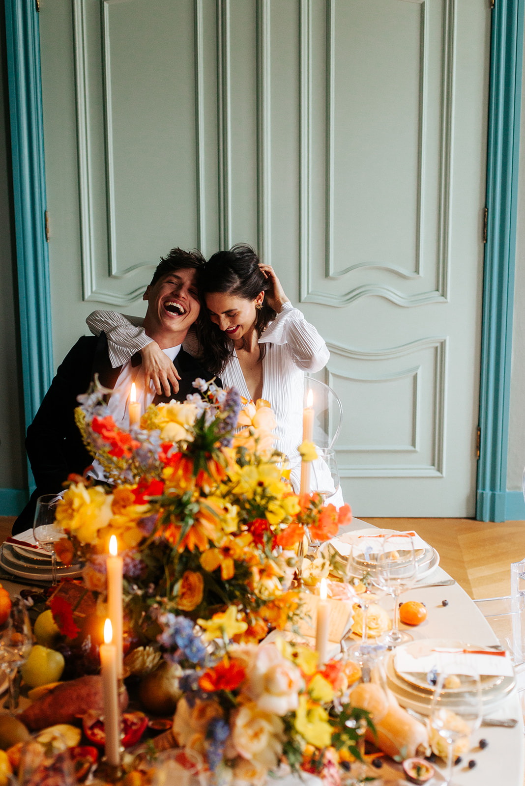 Zeer kleurrijke afbeelding, met oranje, gele, rode en blauwe bloemen die in een prachtig boeket op de tafel staan. De tafel is netjes gedekt en er staan kaarsen op tafel. Verder is de tafel versierd met fruit. Twee gelukkige mensen omhelzen elkaar en lachen, in een keurig onderhouden, nette kamer. De stijl van het huis lijkt een oud landhuis.