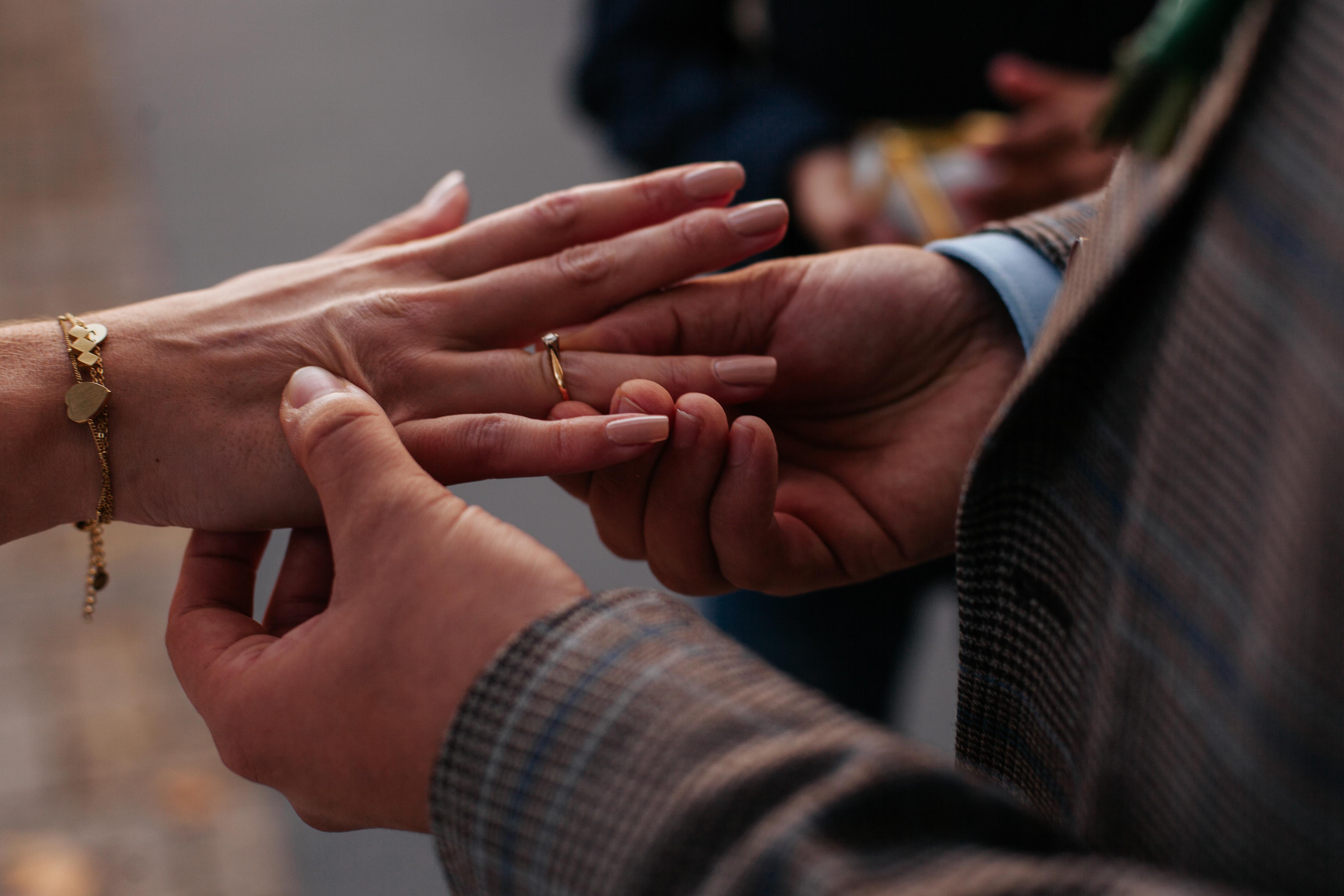 Een vrouwenhand die wordt vastgehouden door de twee handen van een man. Met een van zijn handen schuift hij de huwelijksring om haar ringvinger.