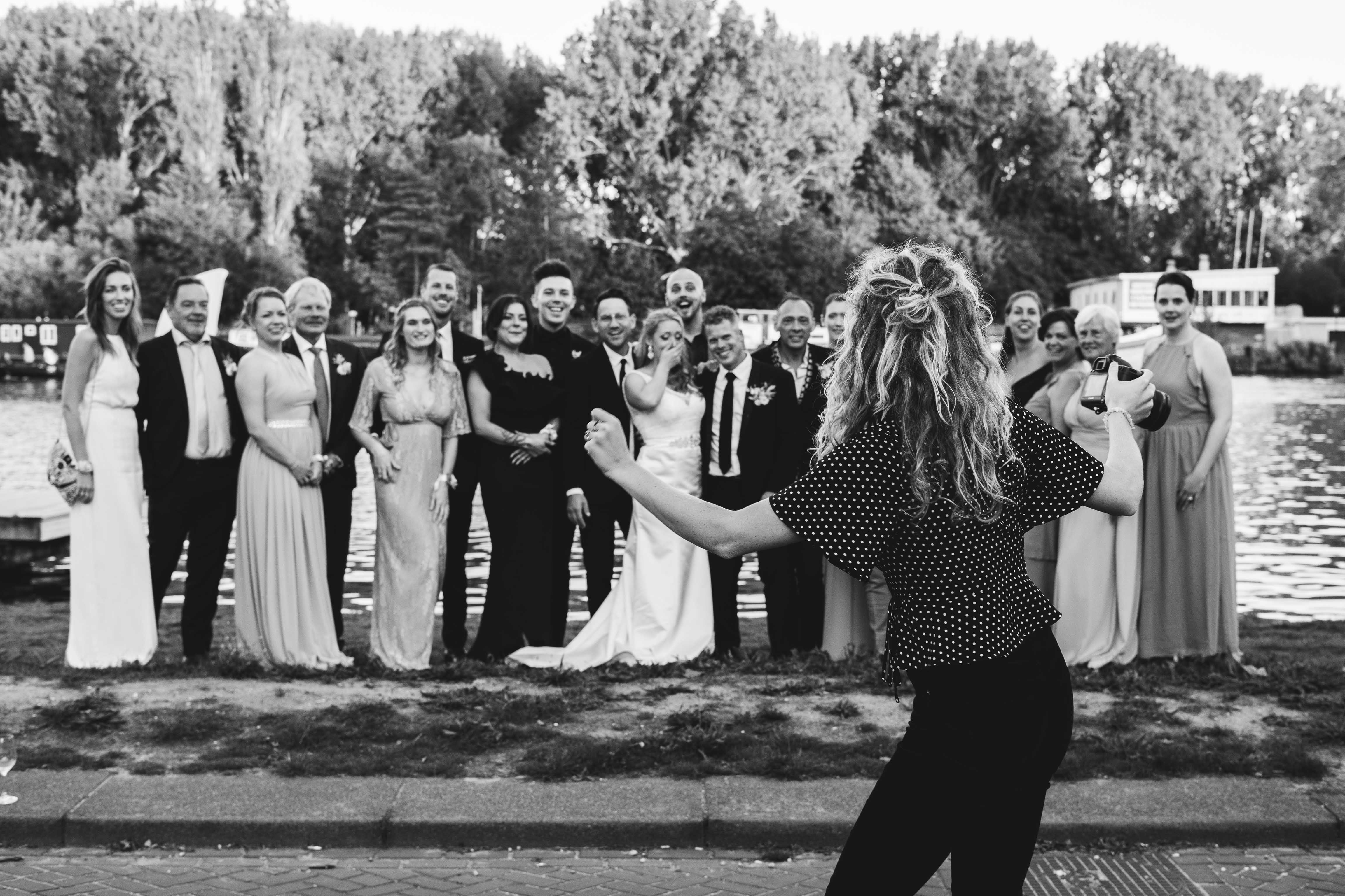 Yara Photography geeft instructies aan een groep mensen voor een groepsfoto. De mensen, allemaal in pak en jurk gekleed, staan voor het water bij elkaar om samen op de foto te gaan.