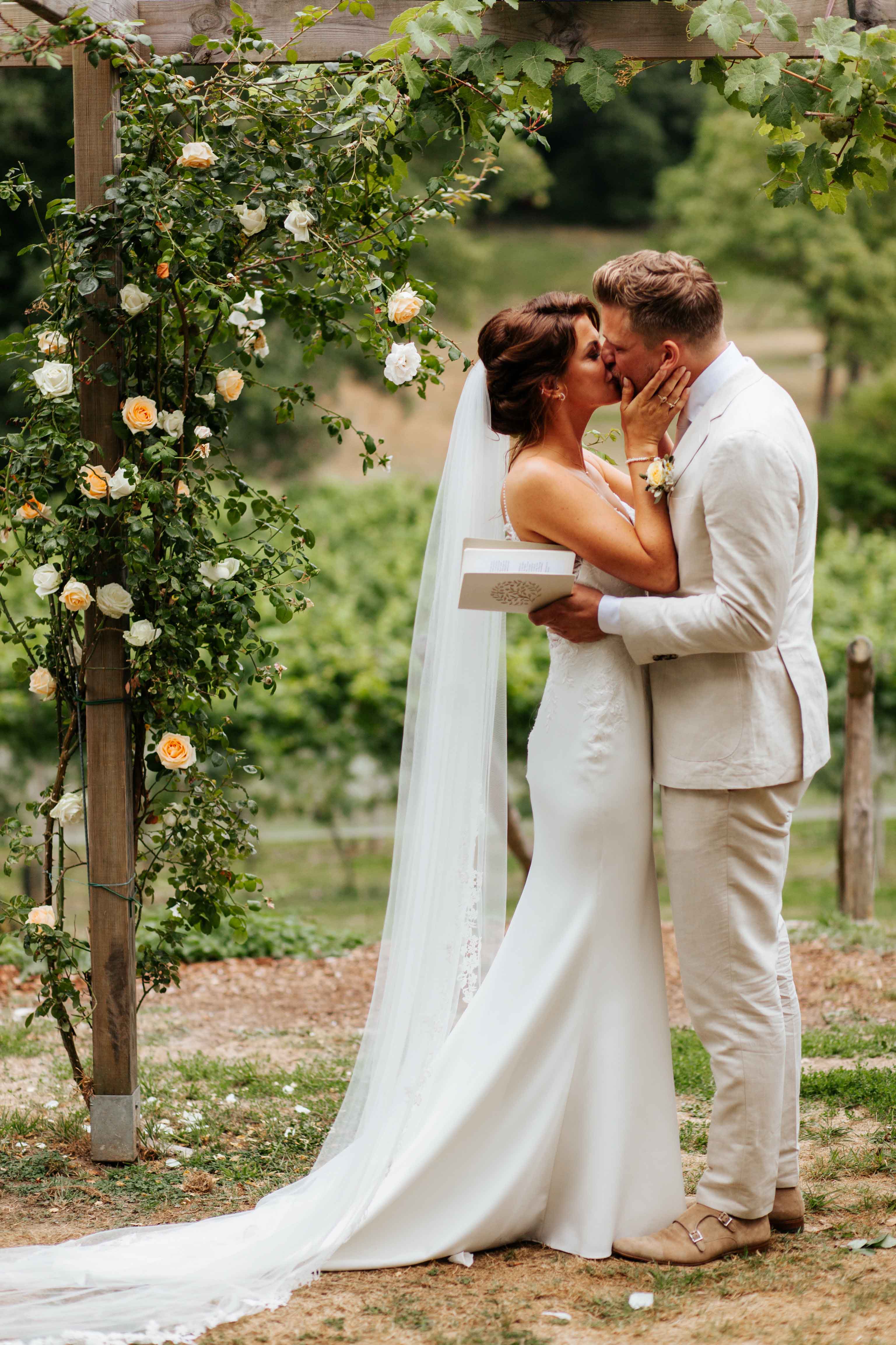 Een echtpaar kust elkaar bij de ceremonie van hun huwelijk. Beide dragen lichte kleding en in de achtergrond is een wijngaard zichtbaar.