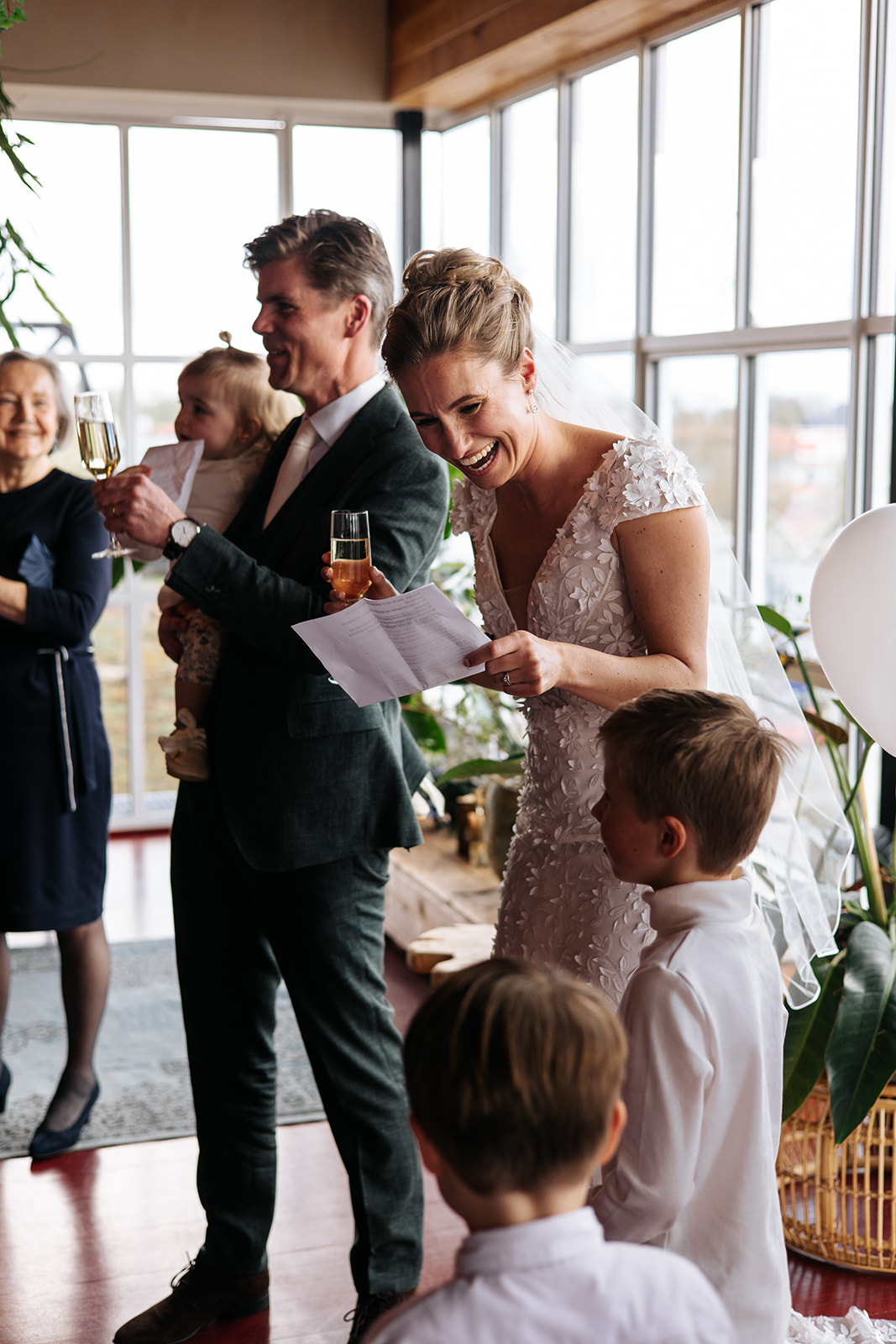 Bruid en bruidegom glimlachen tijdens toast met kinderen in lichte kamer.