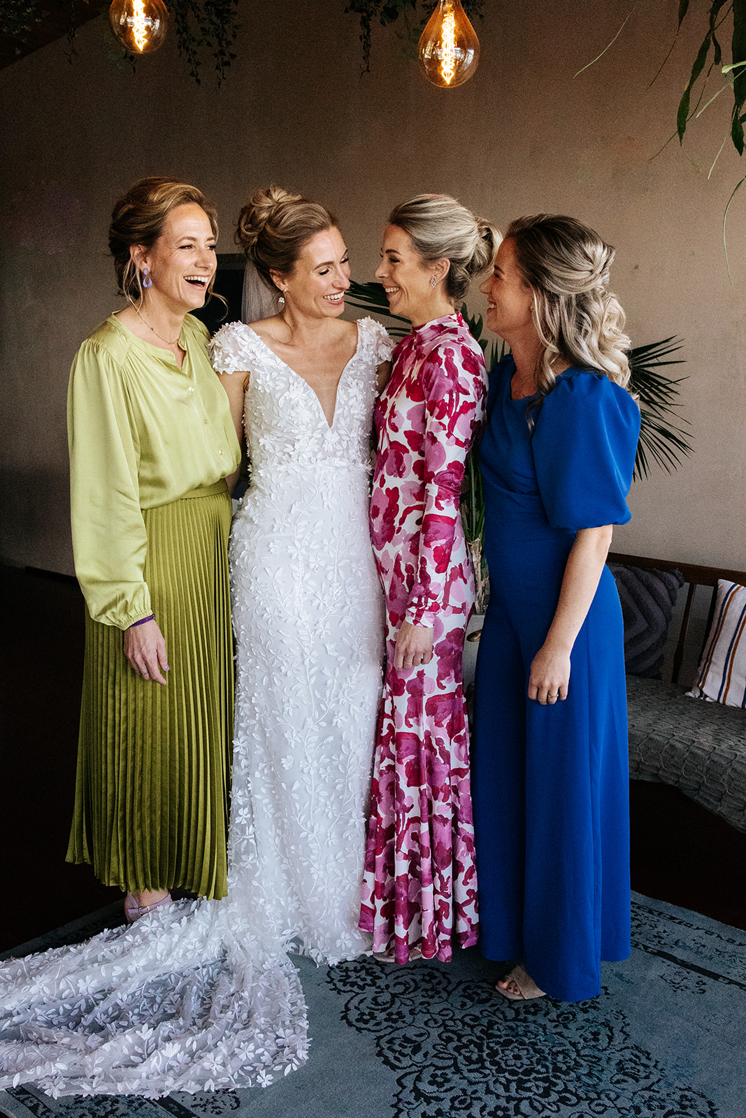 Bruid in witte jurk lacht met drie vrouwen in kleurrijke jurken, gezellig samen staand.
