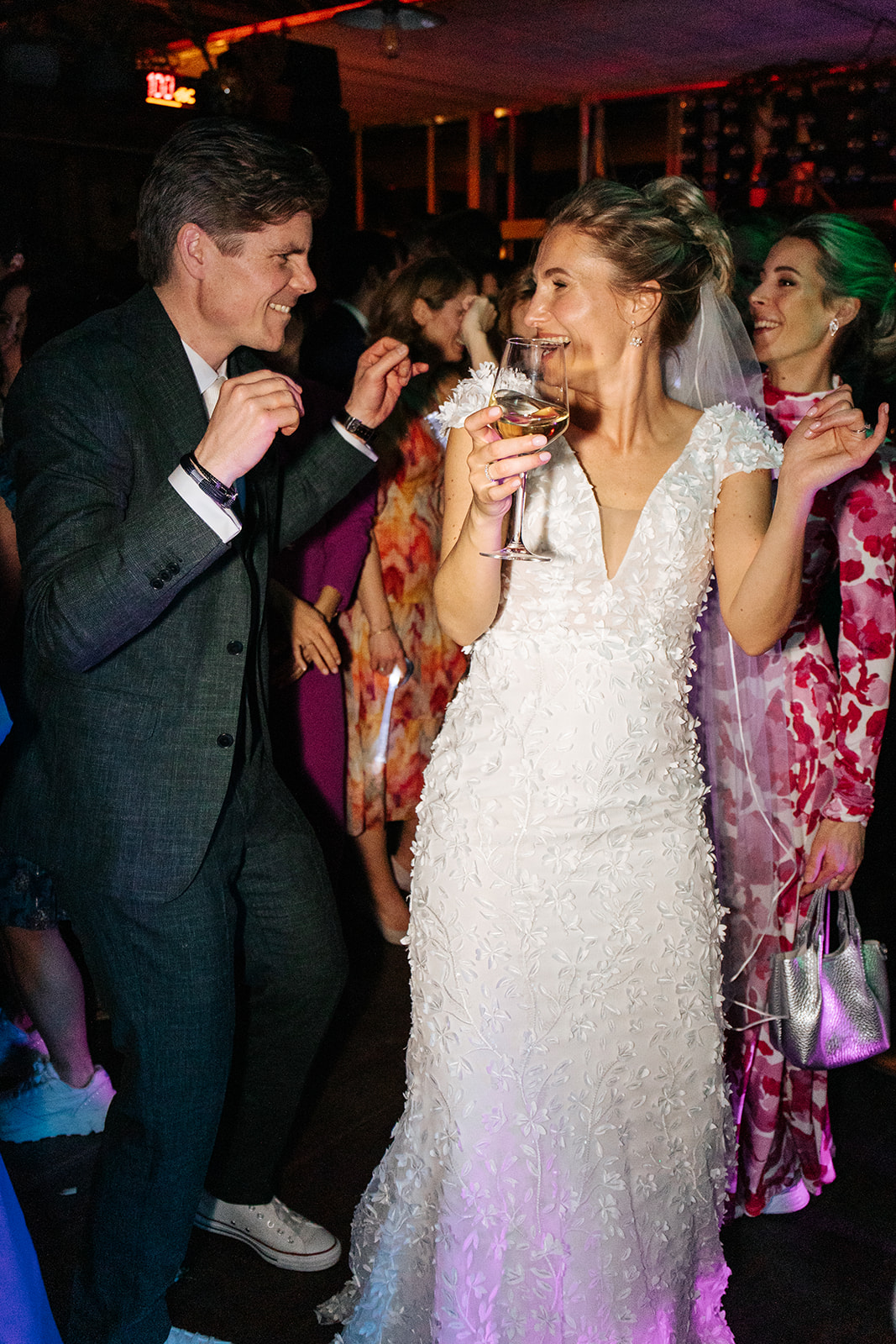 Bruid in witte jurk danst en lacht met man in pak op feest, met glas wijn in haar hand.