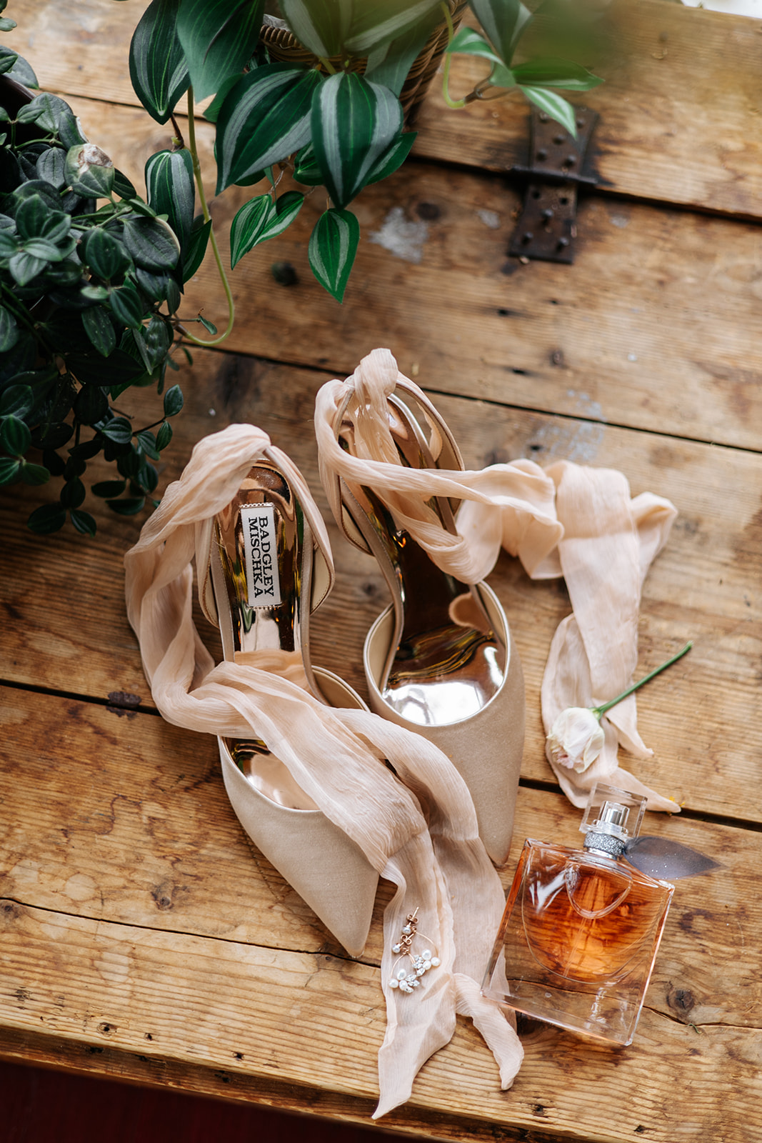 Bruidsaccessoires op een houten tafel: beige schoenen met linten, oorbellen en een parfumfles naast groene planten.