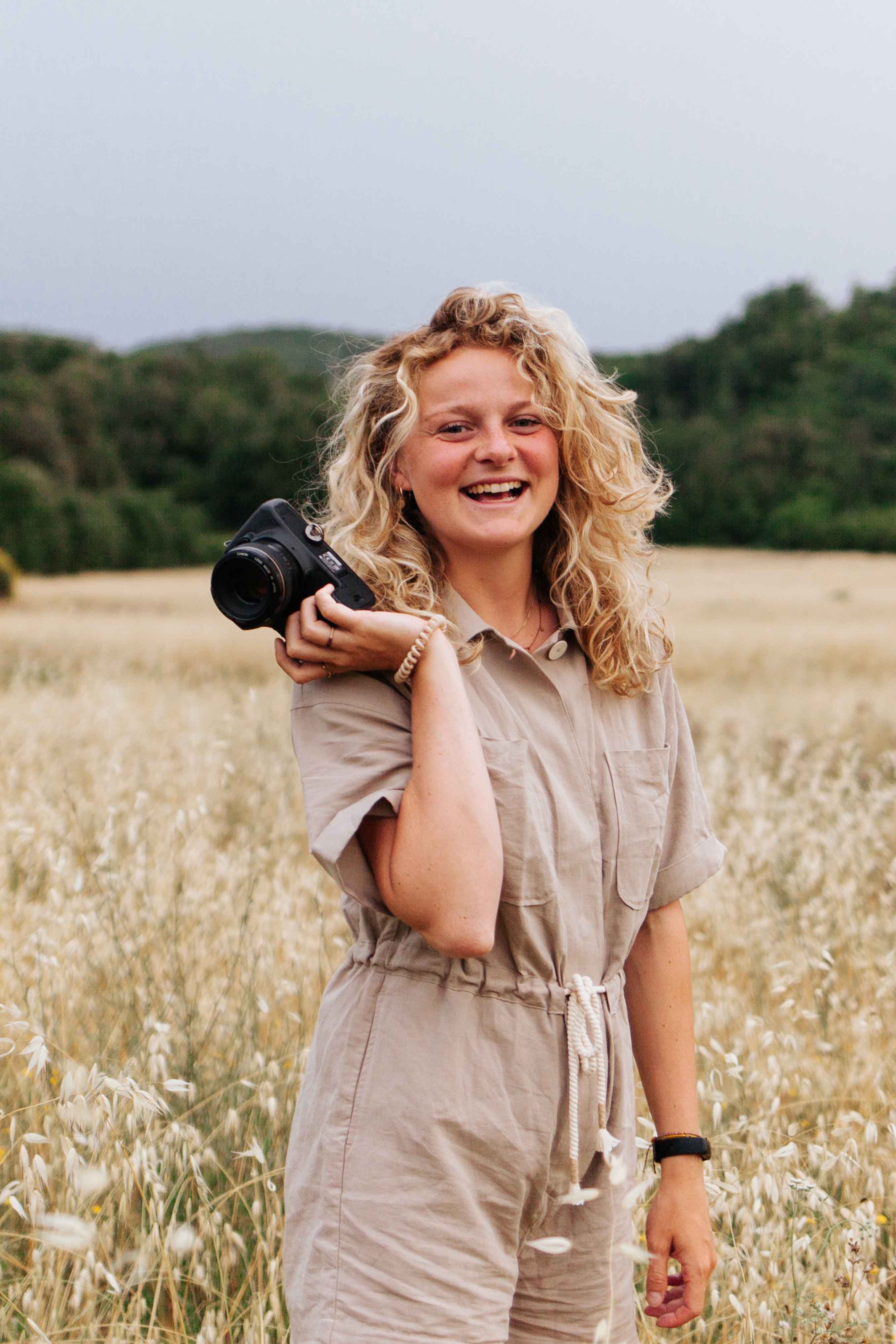 De fotografe van Yara Photography staat in een weiland, waar droog gras te zien is in de achtergrond. Zij poseert voor de camera, met een camera in haar hand en lacht breed. Ze draagt een beige jumpsuit en haar blonde krullen hangen los langs haar hoofd.