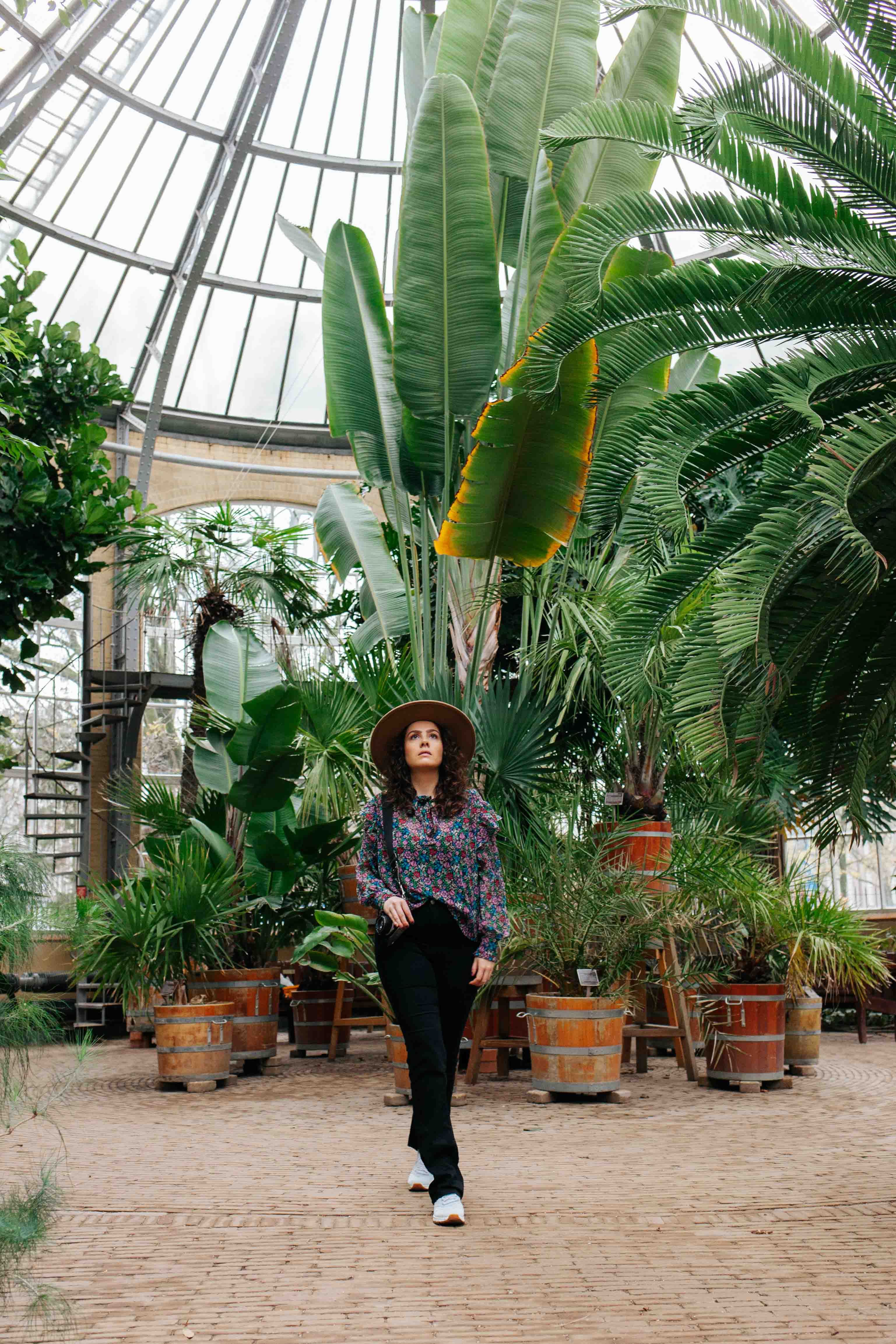 Fleur van Fris Film poseert in de Hortus Botanicus in Amsterdam. Zij draagt een zwarte broek, lichte sneakers en een gekleurd bloemenbloesje. Zij staat in een grote kas, met tropische planten in de achtergrond.